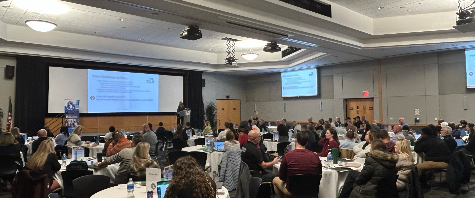 A conference room filled with attendees seated at round tables, watching a presentation on a large screen.