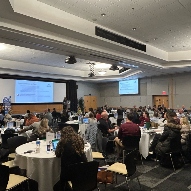 A conference room filled with attendees seated at round tables, watching a presentation on a large screen.