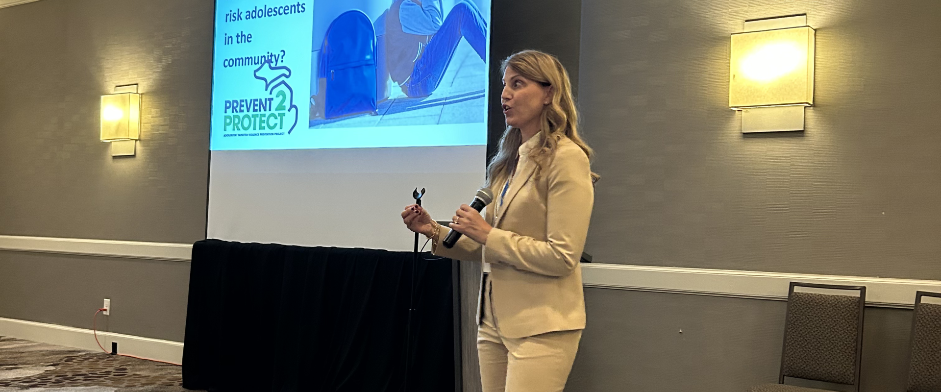 A woman in a beige suit presents in a conference room, with a slide about managing high-risk adolescents projected behind her.