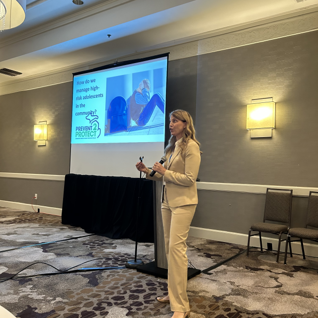 A woman in a beige suit presents in a conference room, with a slide about managing high-risk adolescents projected behind her.