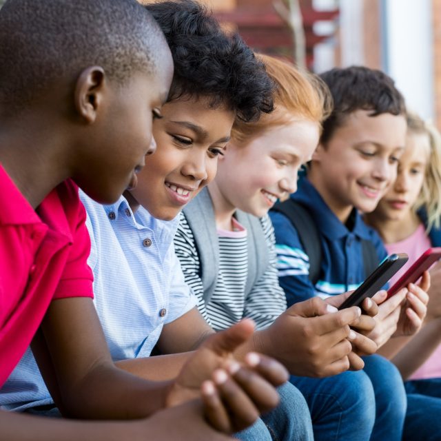 Children sitting outdoors, looking at mobile phones.
