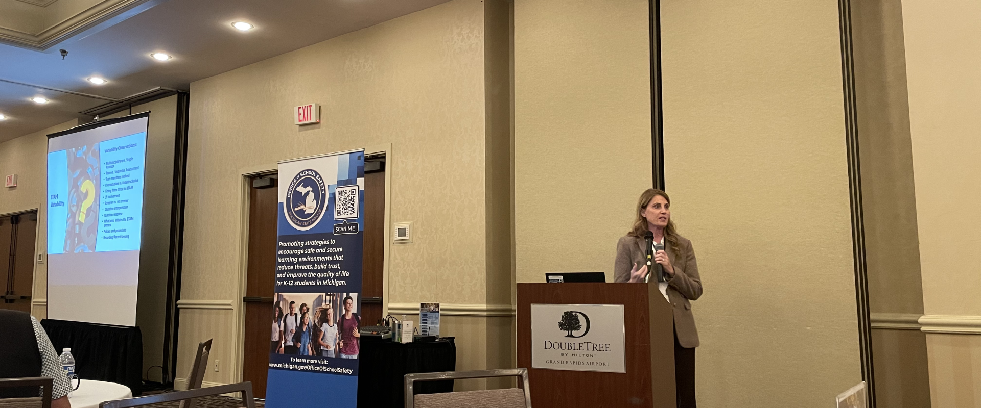 Woman presenting at a podium with a projected slide and banner in a conference room.