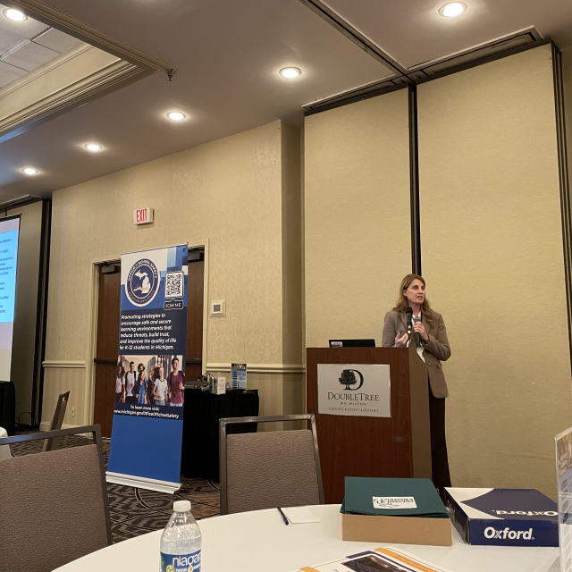 Woman presenting at a podium with a projected slide and banner in a conference room.