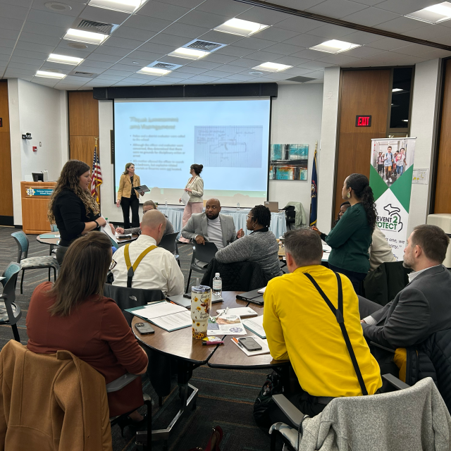 A conference room with people attending a presentation. A screen displays a slide, surrounded by attendees seated and standing.